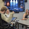 3 people at a table laughing and playing a game. One has a sticker-covered laptop open. One is using a wheelchair and a speech-generating device. 
