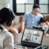professional woman wearing headphones attends meeting on laptop at home with family nearby